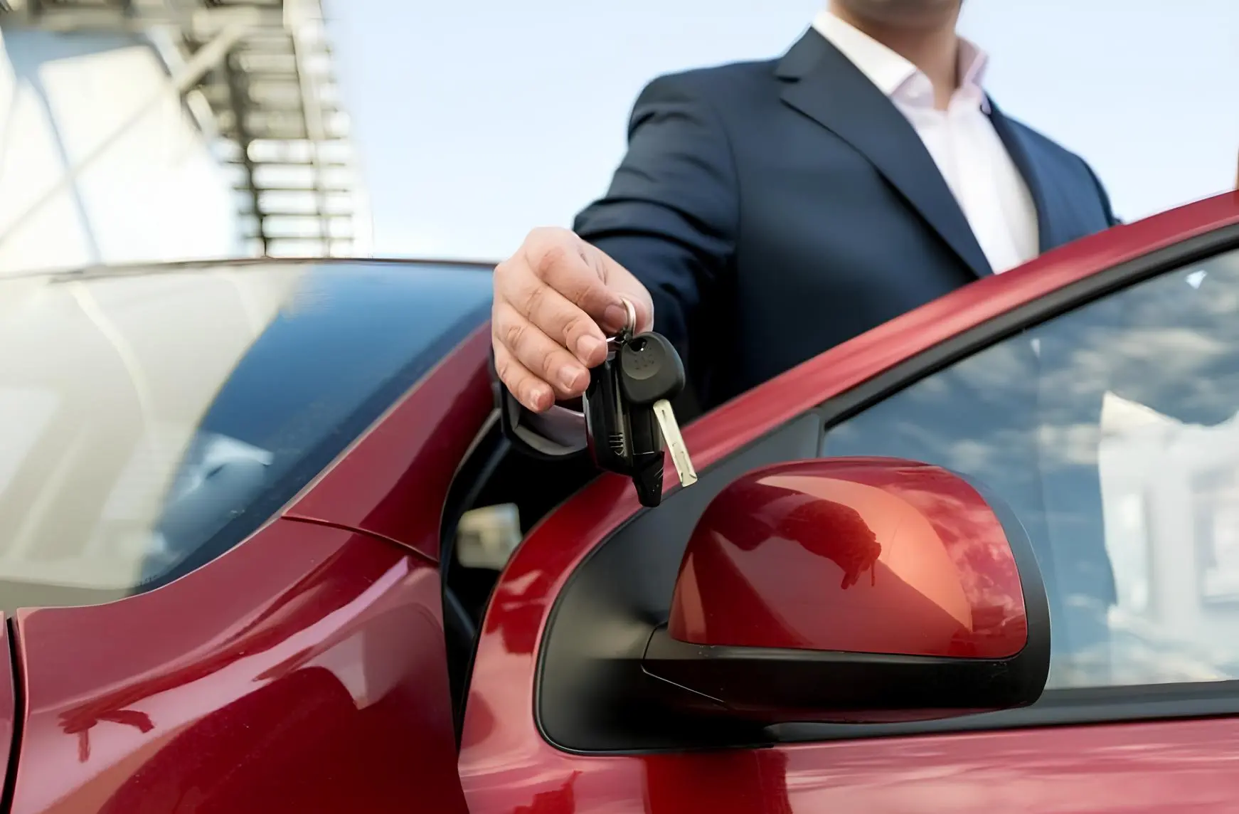 Чоловік у діловому костюмі простягає ключі від автомобіля біля червоного авто на відкритій парков