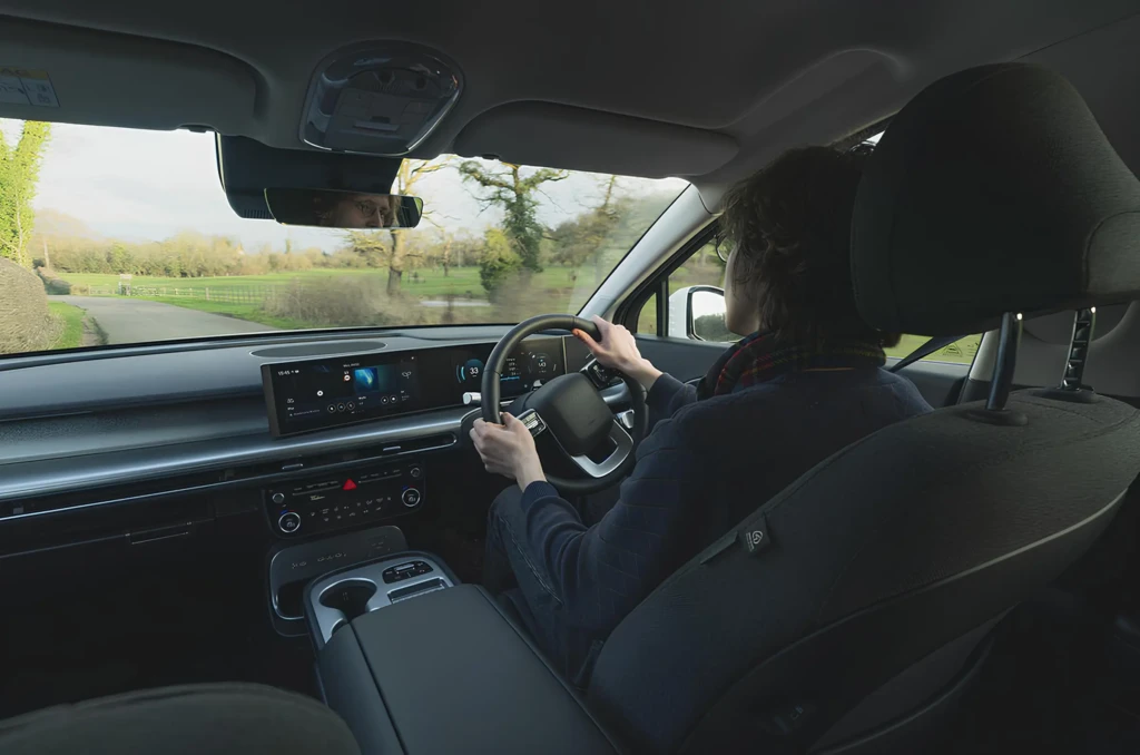 Вид із другого ряду на салон авто під час руху: водій тримає кермо, попереду широка панель приладів із центральним екраном, за вікном — заміська дорога та зелений пейзаж.