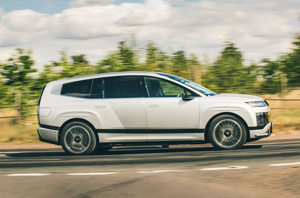 Білий електричний кросовер Hyundai Ioniq 9 у профіль під час руху шосе: довгий кузов, затемнені вікна, великі колеса, розмитий зелений фон.