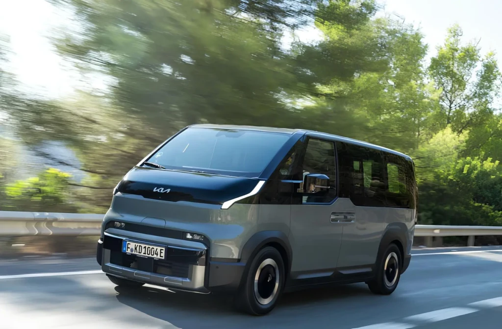 Електричний мінівен Kia їде шосе: двоколірний кузов, великі вікна, розмитий зелений фон лісу.