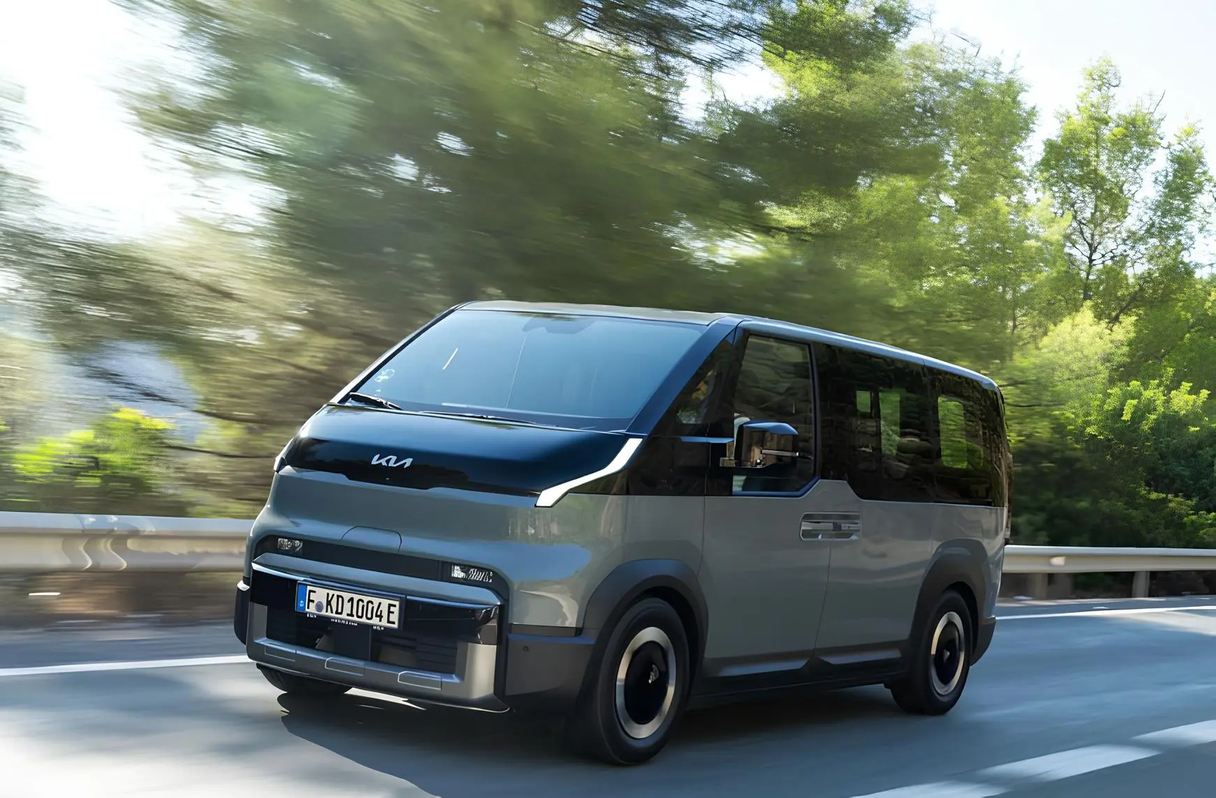 Електричний мінівен Kia їде шосе: двоколірний кузов, великі вікна, розмитий зелений фон лісу.