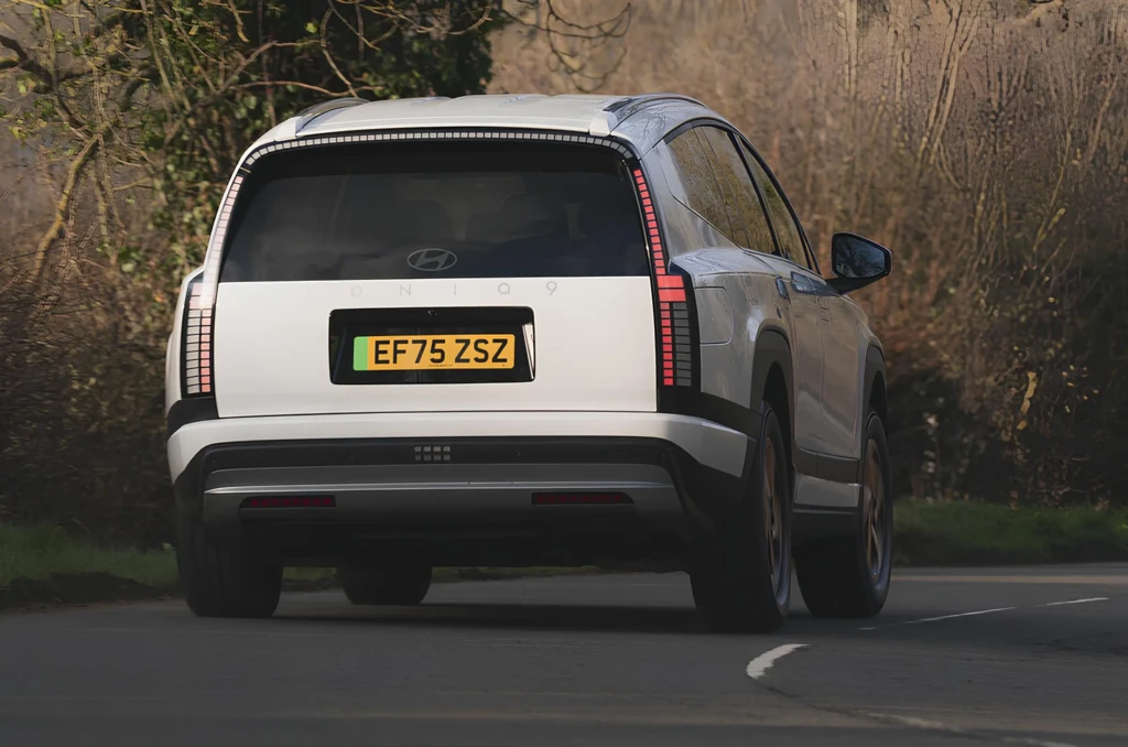 Задний ракурс белого электрического кроссовера Hyundai Ioniq 9 в движении на извилистой дороге: вертикальные LED-фонари, надпись IONIQ 9 на крышке багажника, лесной фон.