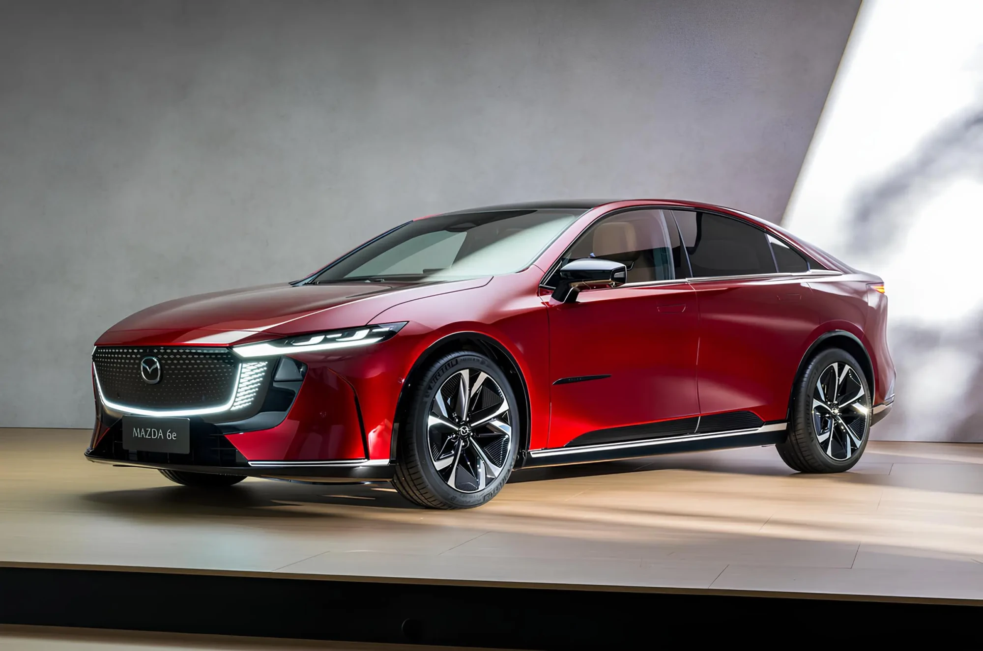 Червоний седан Mazda 6e на презентаційному подіумі: тричвертний ракурс спереду, світлодіодна оптика та підсвічена решітка радіатора Mazda