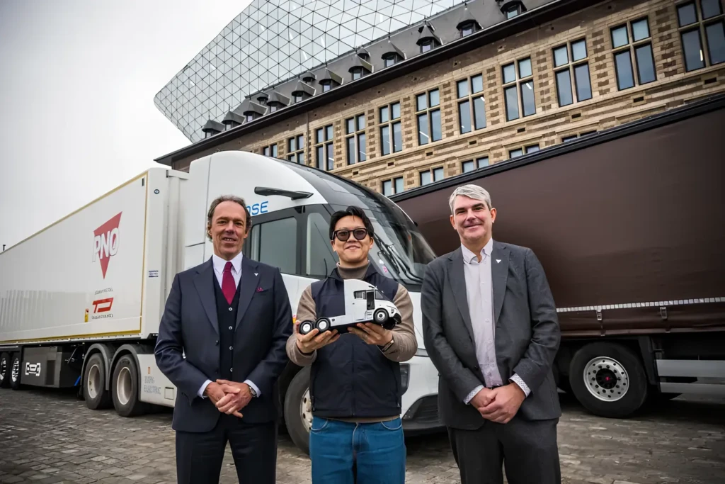 Троє людей позують біля білого електричного тягача з напівпричепом. Людина по центру тримає модель вантажівки.