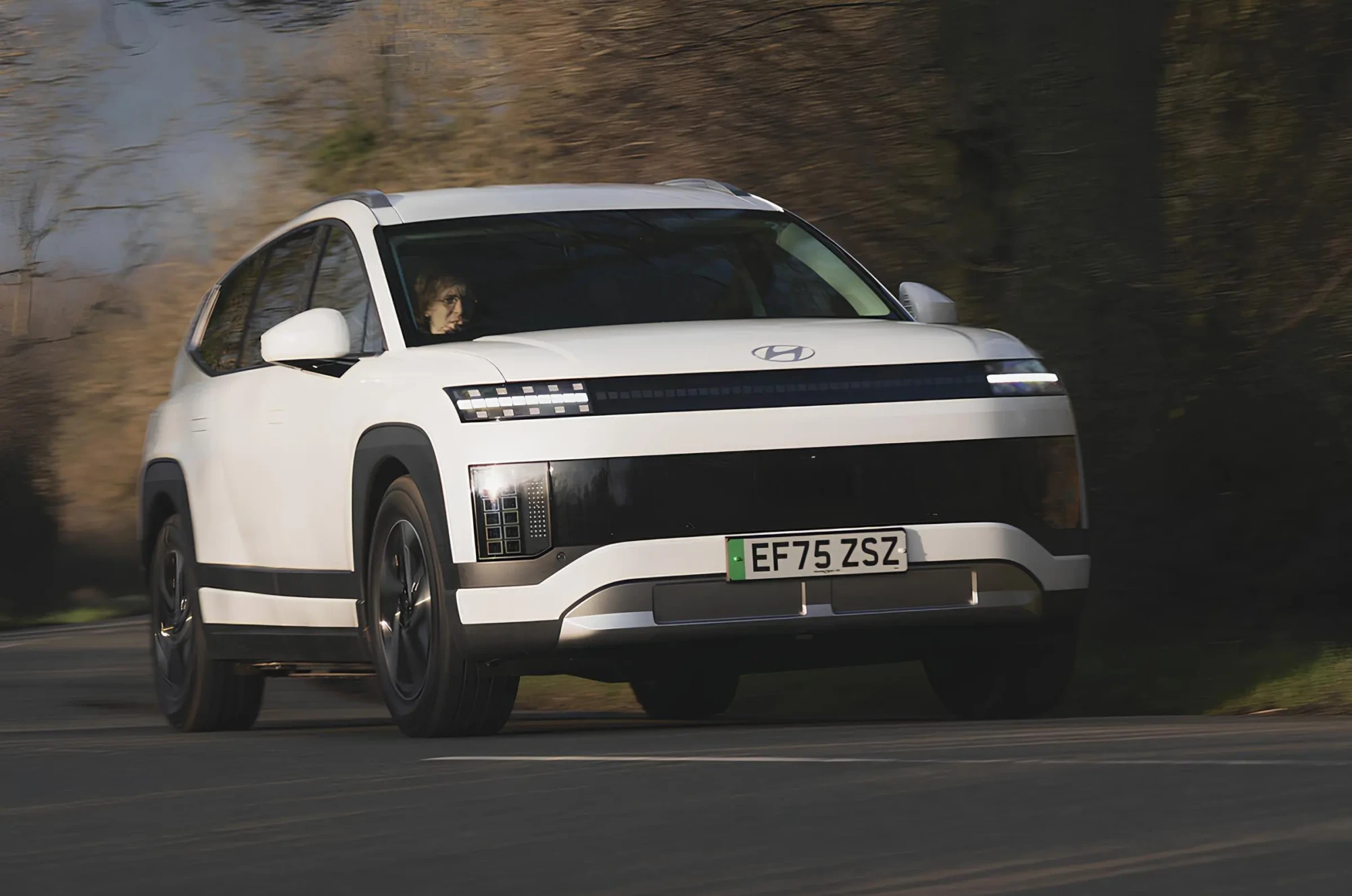 Білий електричний кросовер Hyundai у русі на дорозі: фронтально-боковий ракурс, піксельна LED-оптика, розмитий фон дерев.