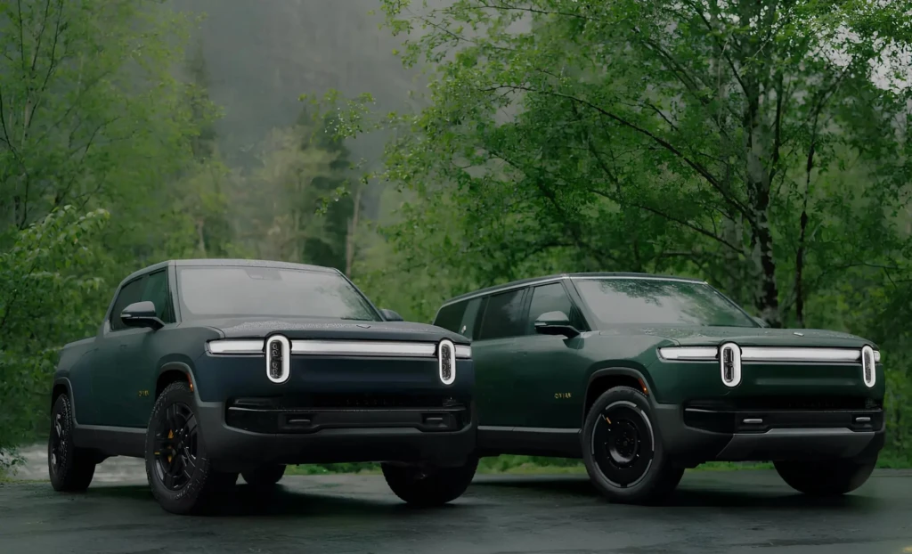 Два електричні пікапи Rivian Dual Standard у темно-зеленому кольорі на мокрій дорозі в лісі, фронтальний ракурс із фірмовими вертикальними LED-фарами