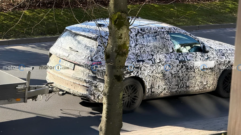 Замаскований прототип Audi Q9 у чорно-білому спіральному камуфляжі, вид ззаду-збоку; кросовер тягне причіп на дорозі, у кадрі частково дерево
