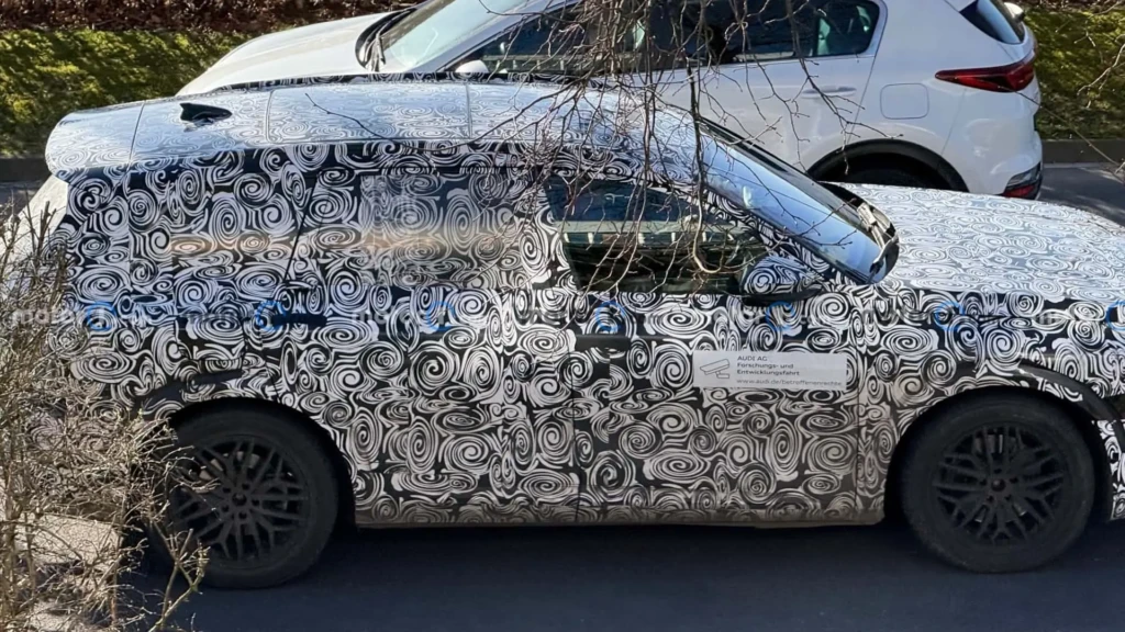 Замаскований прототип Audi Q9 у камуфляжі з біло-чорними спіралями, вид збоку; великий кросовер на парковці поруч з іншими авто