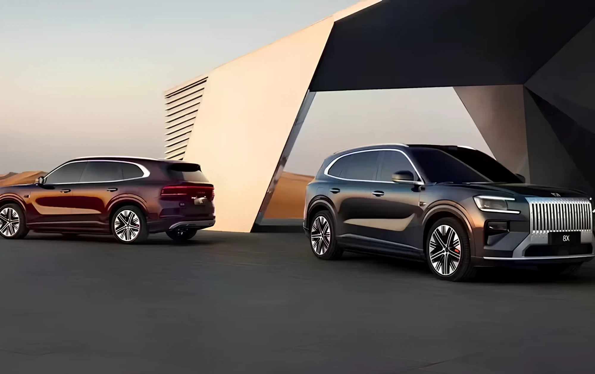 Два кросовери Zeekr 8X біля футуристичної будівлі на тлі пустельного пейзажу: темно-сірий спереду, бордовий позаду