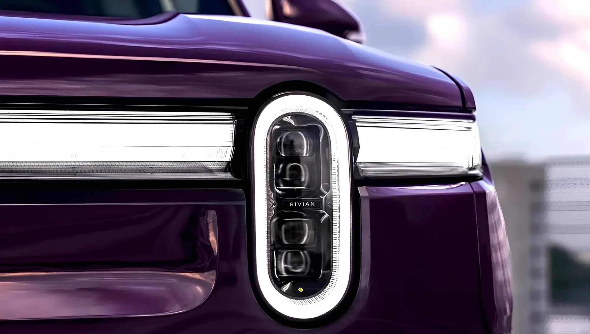 Крупний план передньої оптики Rivian Dual Standard: овальна вертикальна LED-фара з трьома модулями та горизонтальна світлова смуга на фіолетовому кузові