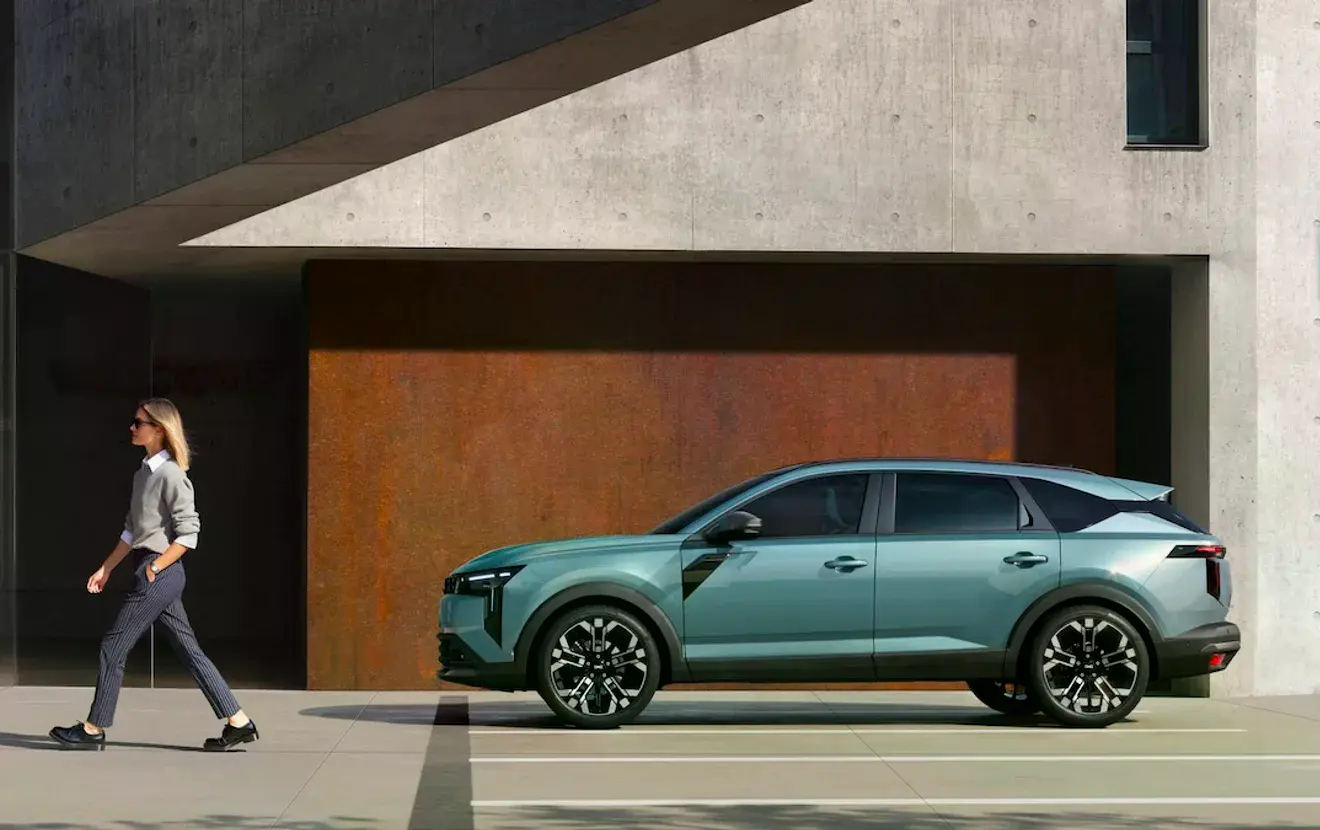 Dacia Striker: бірюзовий кросовер у профіль біля сучасної бетонної будівлі, поруч проходить жінка