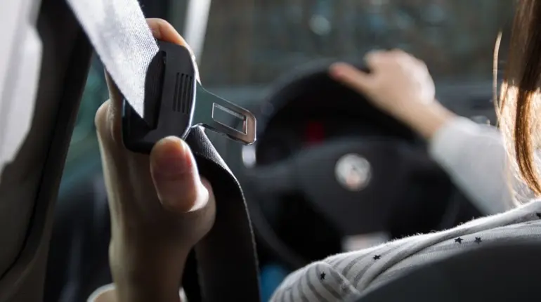 Водій у салоні авто тримає ремінь безпеки перед пристібанням; на фоні видно кермо та панель приладів у розфокусі