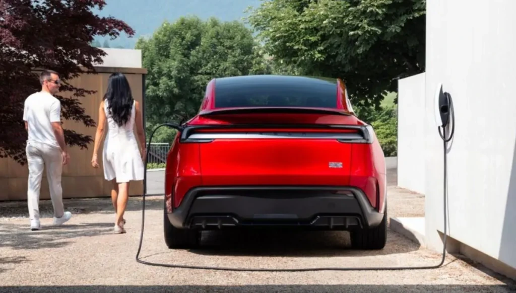 Tesla Model 2 червоного кольору під час заряджання біля будинку, вигляд ззаду